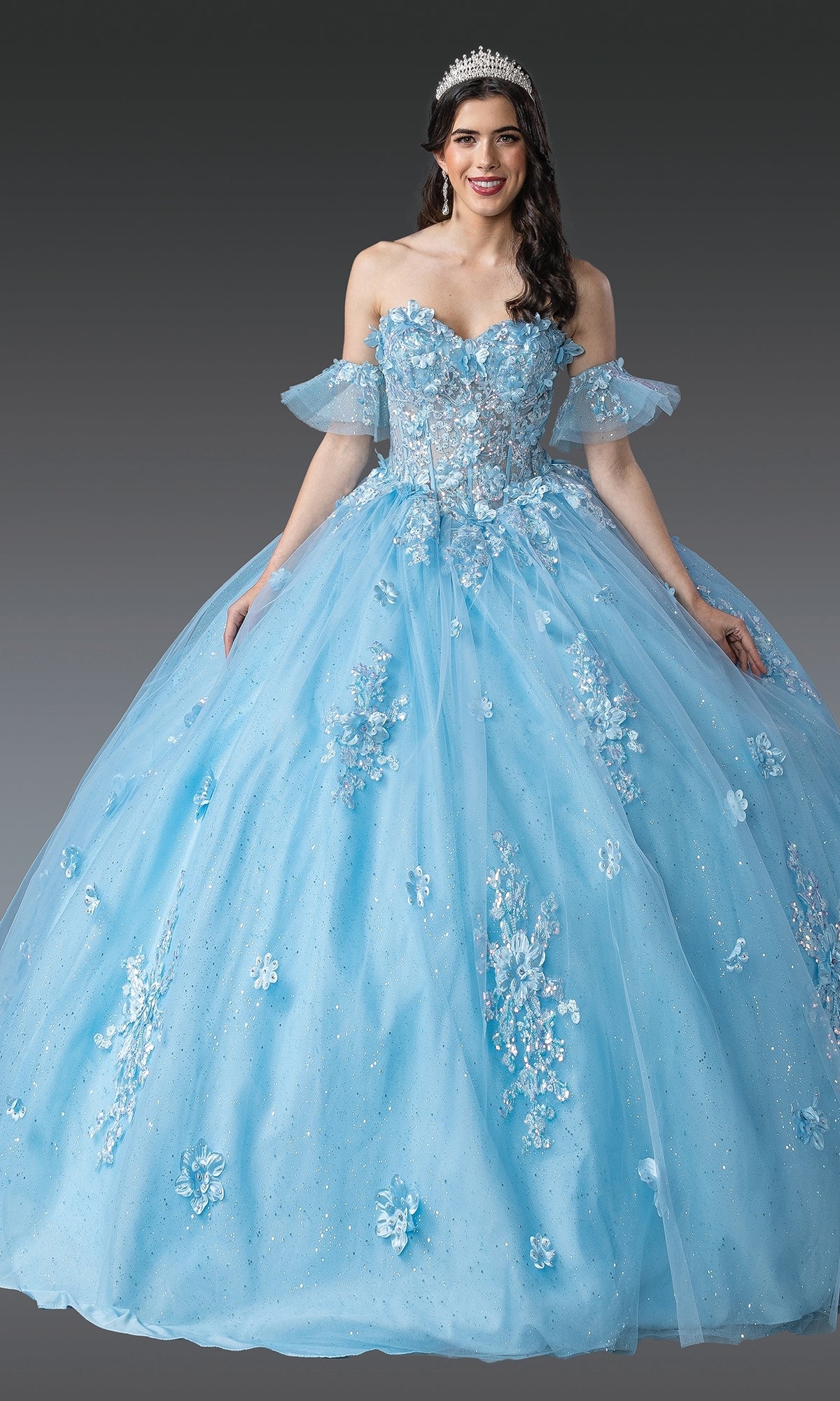 A woman poses against a plain gray background wearing the Blue Quinceanera Dress 1964 By Dancing Queen, featuring floral embellishments, off-the-shoulder sleeves, and a corset bodice, accessorized with a tiara.
